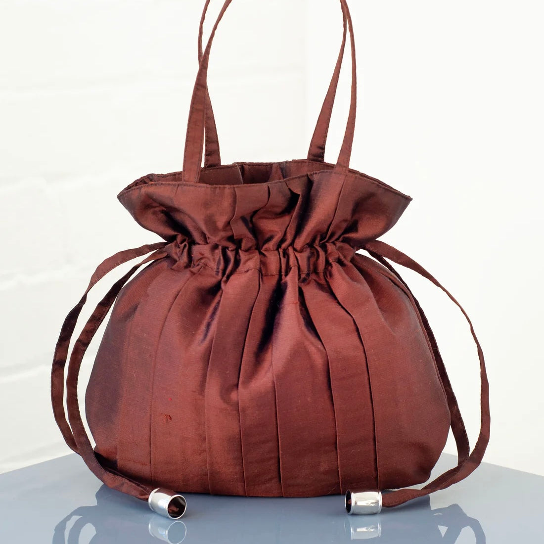 Brown silk drawstring handbag with a top handle and pleats sits on a clear surface with a white background