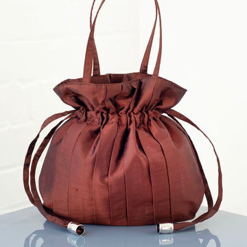 Brown silk drawstring handbag with a top handle and pleats sits on a clear surface with a white background
