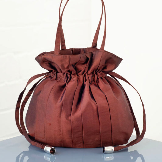 Brown silk drawstring handbag with a top handle and pleats sits on a clear surface with a white background