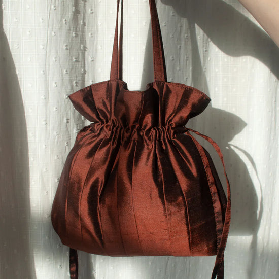 Brown silk drawstring bag held by a hand against a white curtain background