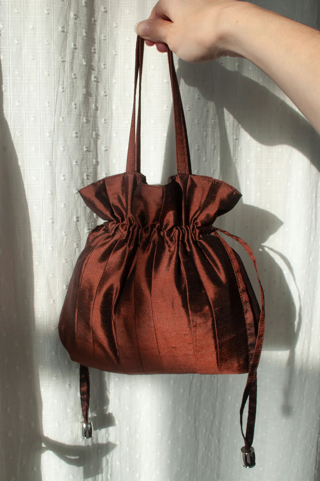 Brown silk drawstring bag held by a hand against a white curtain background