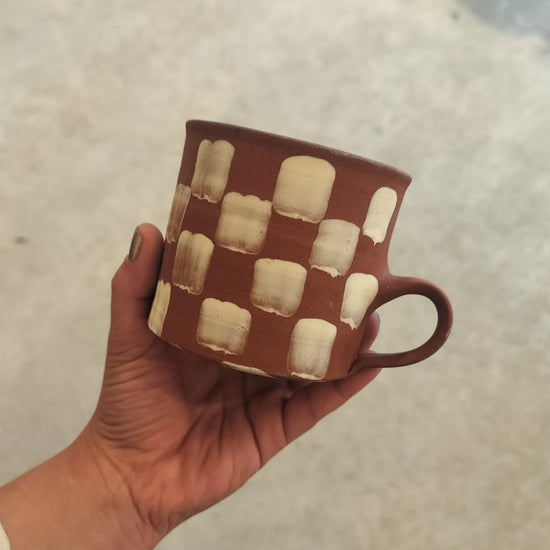 Hand holding a brown ceramic mug with white checkered pattern against a neutral background
