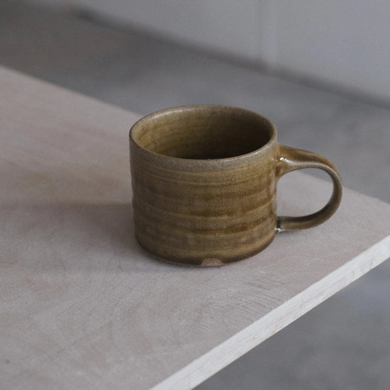 A handmade ceramic mug with a textured, ribbed surface and a warm olive-brown glaze, sitting on the corner of a light-colored wooden table in a minimalist room.