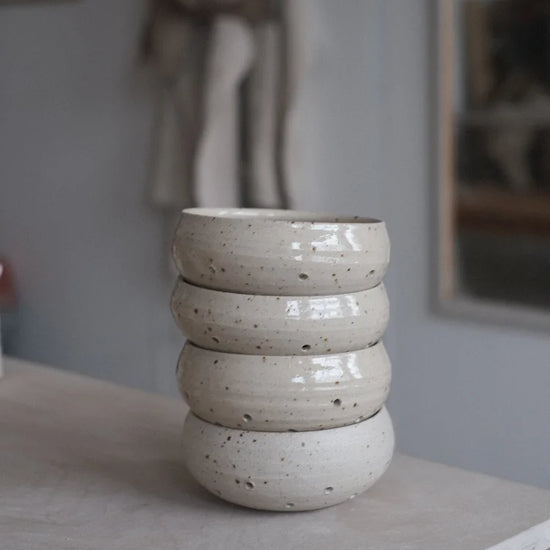 Four grey speckled berry bowls with water drainage holes stacked on top of one another on a table edge on a grey background with an apron hanging up
