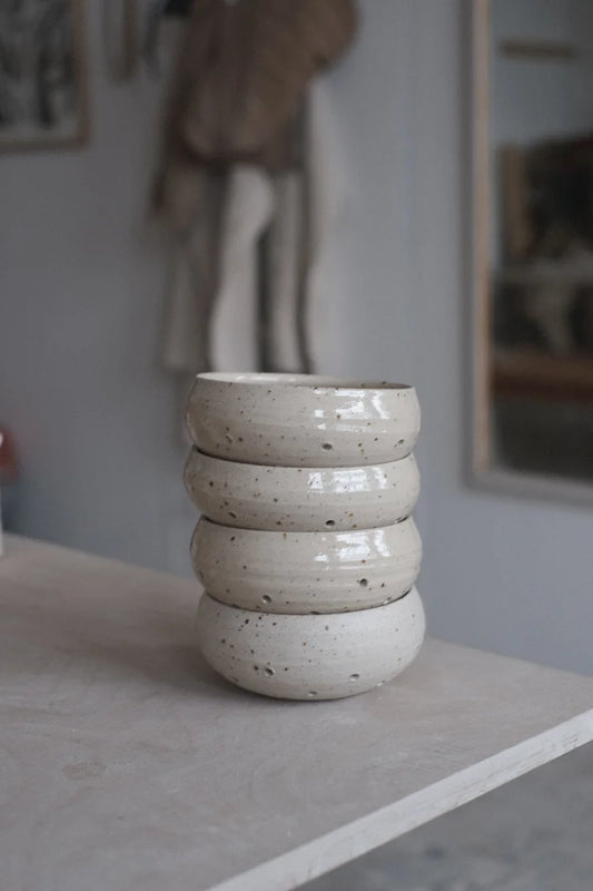 Four grey speckled berry bowls with water drainage holes stacked on top of one another on a table edge on a grey background with an apron hanging up
