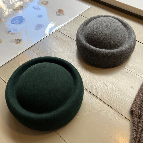 Two wool New York style berets sitting next to each other on a sage green wooden background. One is green and the other is grey
