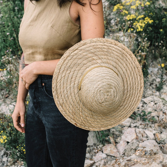 A person holding a beige straw hat with a simple domed crown and a ribbon tie.