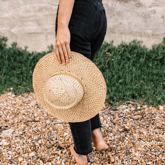 Person holding a straw hat on a pebbly surface with greenery in the background