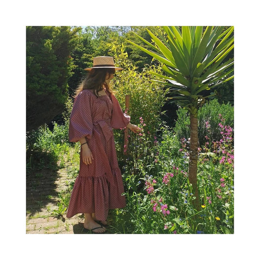 Woman in a pink dress and hat standing in a garden with flowers and trees.