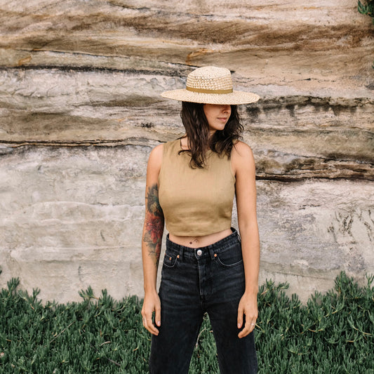 Woman wearing a beige sleeveless top and black pants with a straw hat, standing against a stone wall.