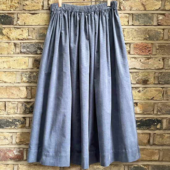 Long pale blue gathered skirt with an elastic waist band on a hanger against a brick wall