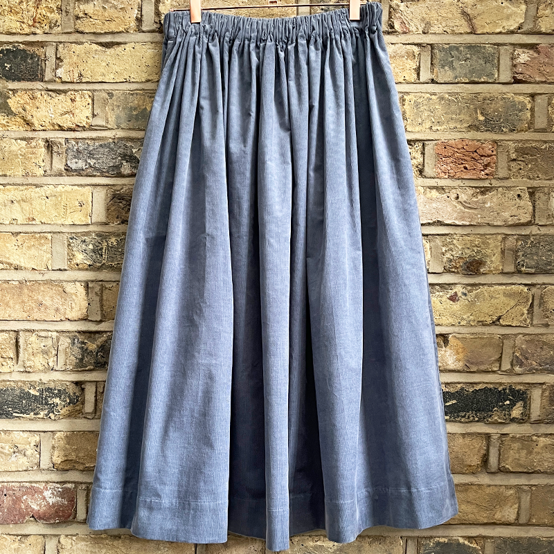 Long pale blue gathered skirt with an elastic waist band on a hanger against a brick wall