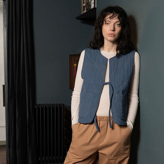 Female model with dark brown hair wears blue padded quilted gilet with a single front tie. The model has a white long sleeved crew neck top on underneath and camel coloured trousers standing against a blue wall in a home