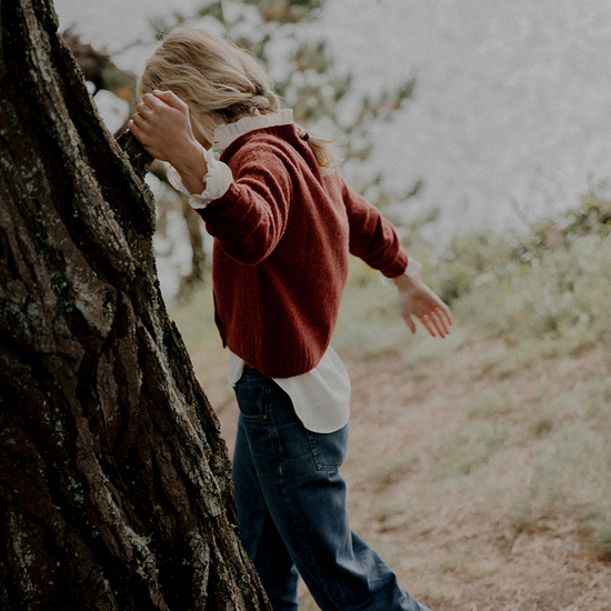 Person in a red sweater and blue jeans standing next to a tree in a natural setting
