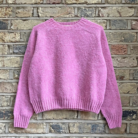 A pink knitted crew neck sweater displayed against a brick wall background.