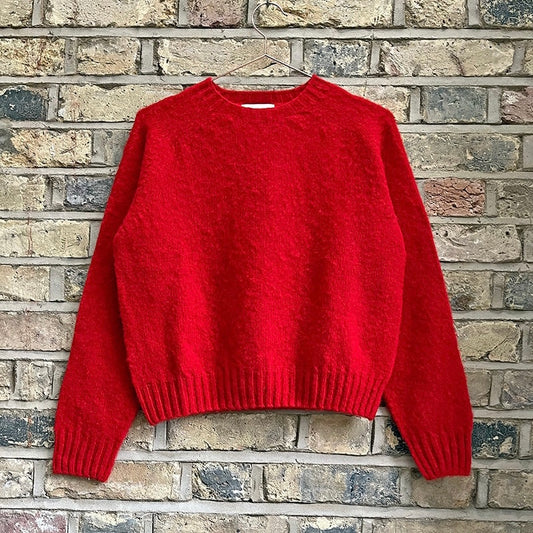 A red knitted sweater with a crew neck and rib knit cuffs and hem, displayed against a brick wall.