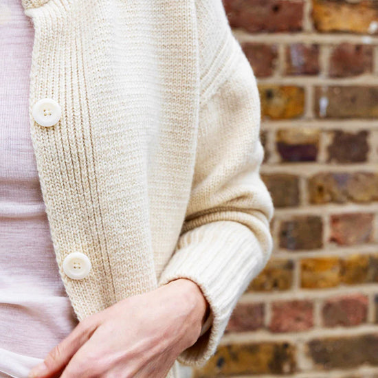 Close-up of a person wearing a cream knitted cardigan against a brick wall.