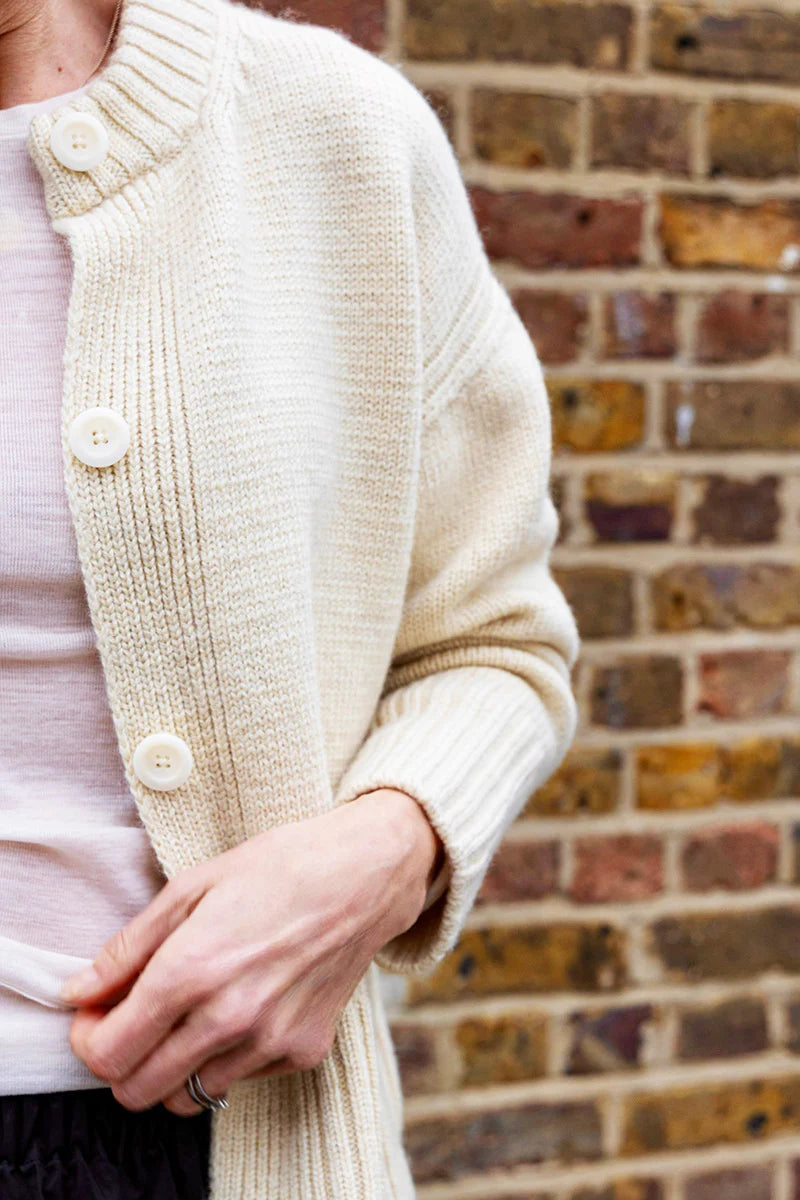 Close-up of a person wearing a cream knitted cardigan against a brick wall.