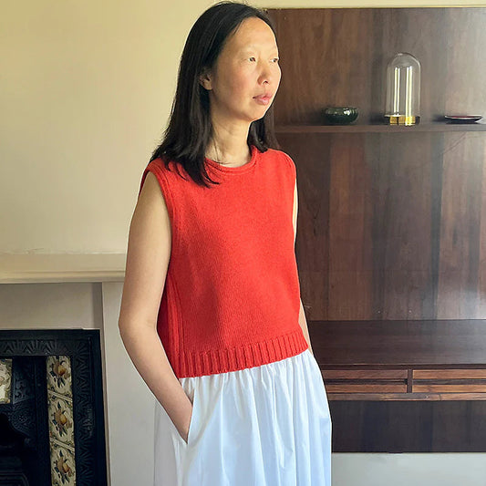 Woman wearing a red sleeveless top and white skirt standing in a room with a wooden cabinet and fireplace.