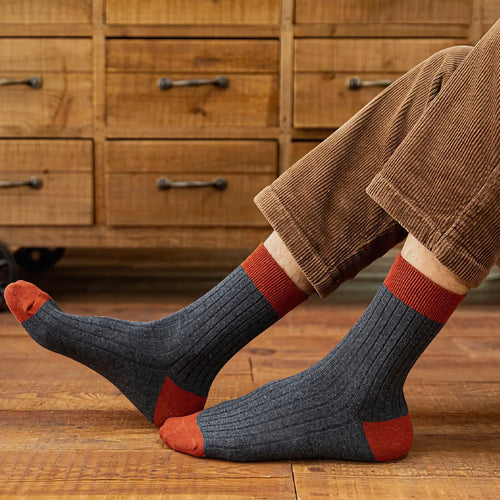 Person wearing grey and red ribbed socks with brown cord trousers in front of wooden furniture and a wooden floor