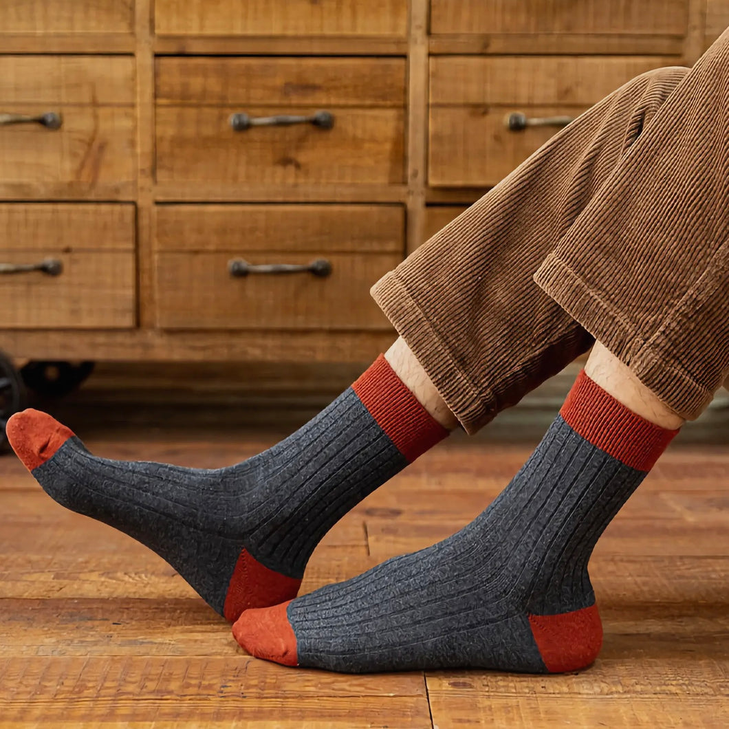 Person wearing grey and red ribbed socks with brown cord trousers in front of wooden furniture and a wooden floor