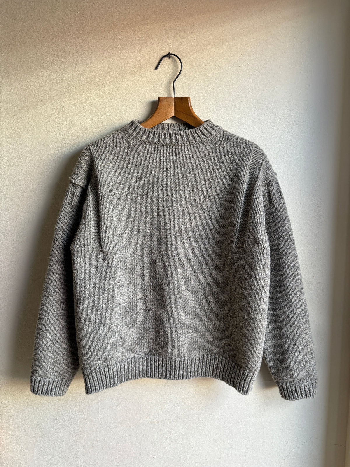 A gray knit sweater hanging on a wooden hanger against a plain off-white wall.
