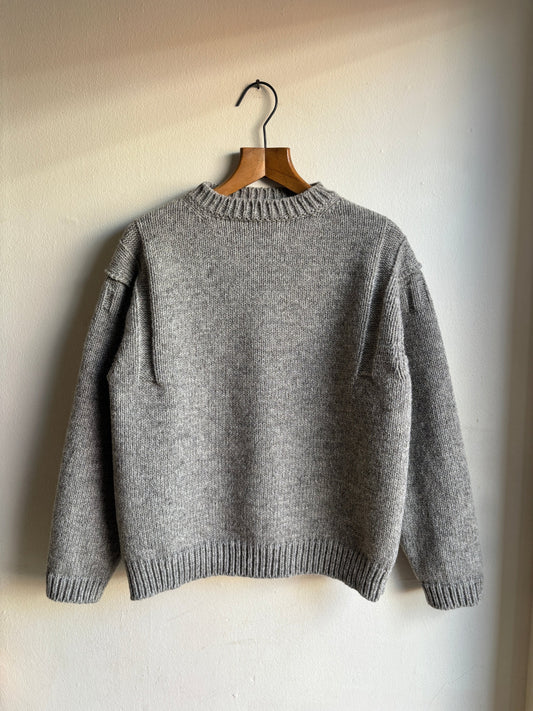 A gray knit sweater hanging on a wooden hanger against a plain off-white wall.