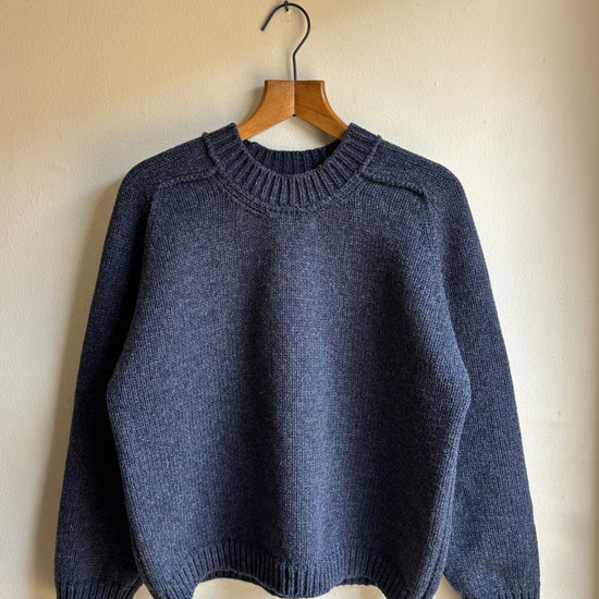 A dark slate blue knit sweater hanging on a wooden hanger against a plain beige wall.