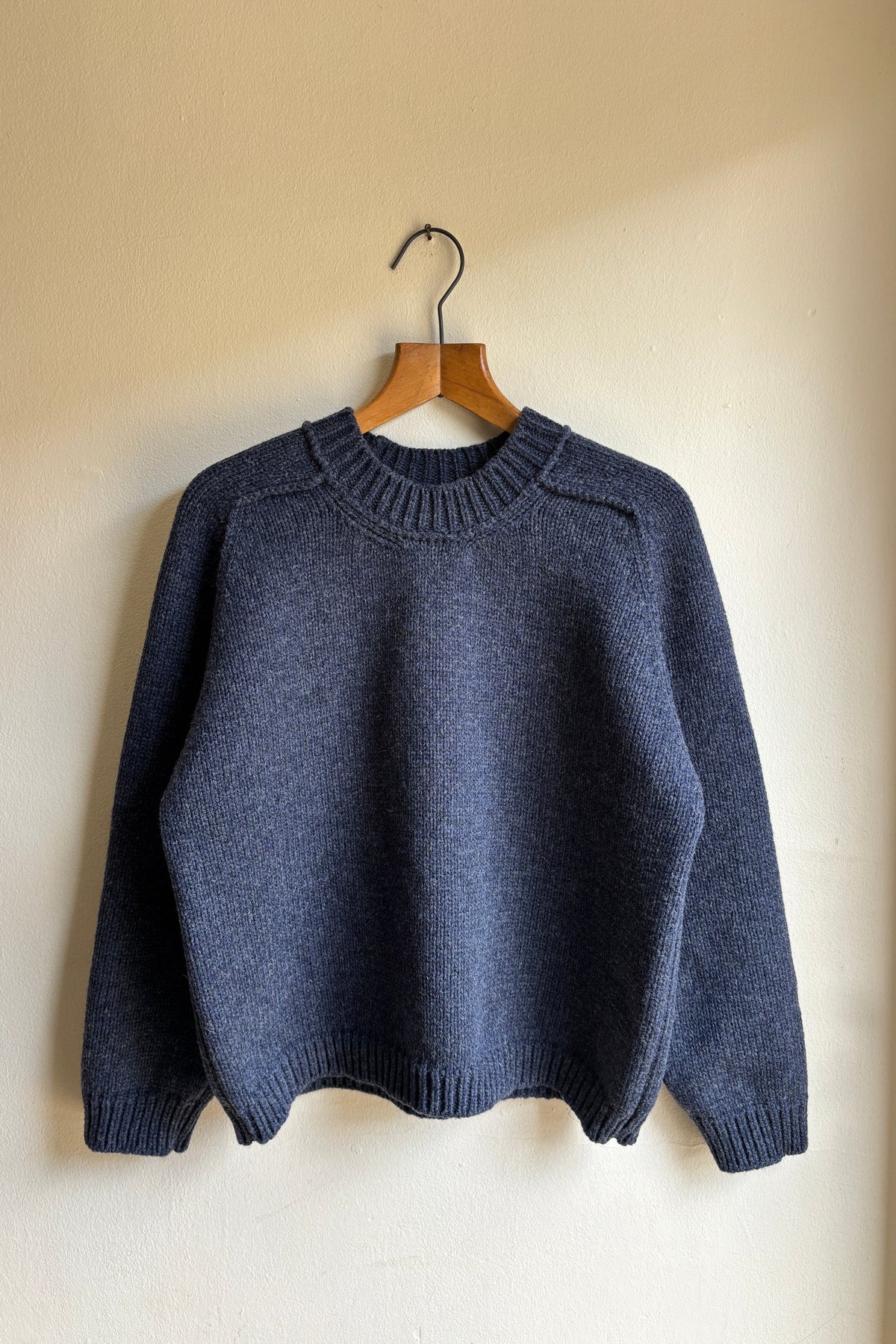 A dark slate blue knit sweater hanging on a wooden hanger against a plain beige wall.