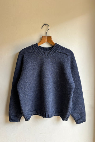 A dark slate blue knit sweater hanging on a wooden hanger against a plain beige wall.