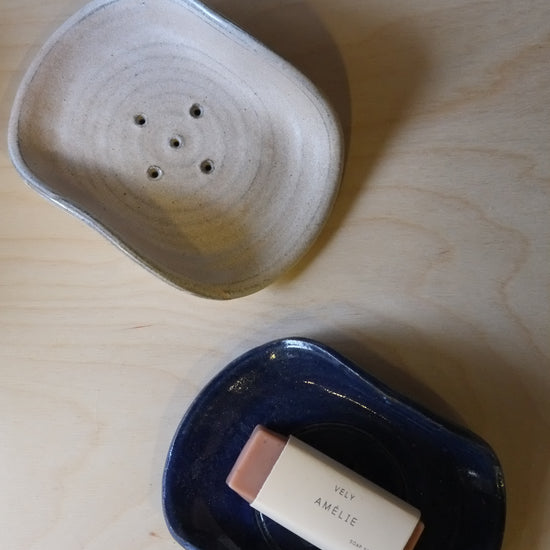 Two ceramic containers on a light wood surface, one with a bar of soap inside.