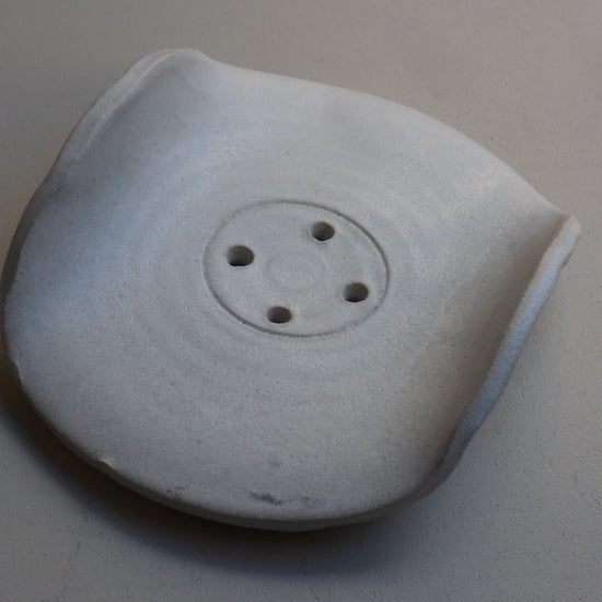 A light grey ceramic soap dish with a circular inset of four small holes, photographed against a neutral light grey background.