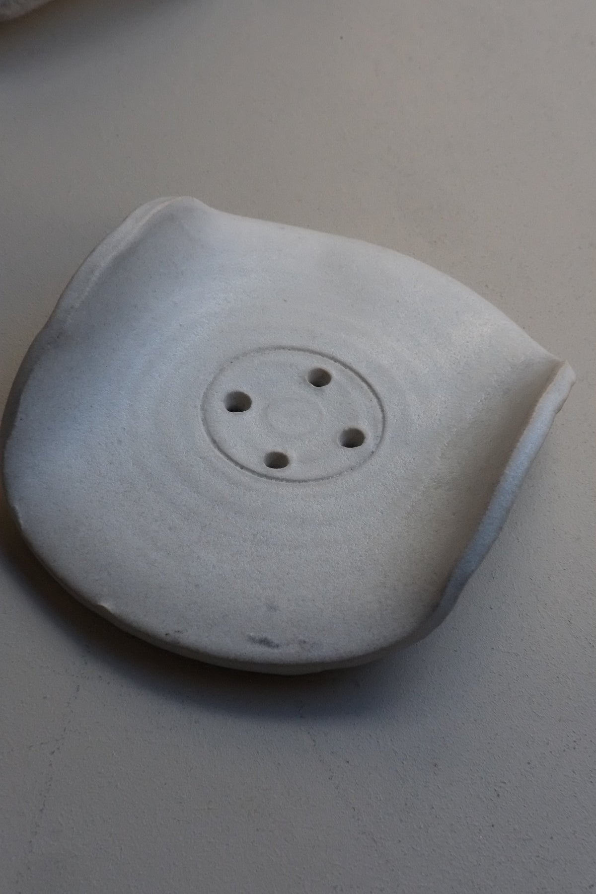 A light grey ceramic soap dish with a circular inset of four small holes, photographed against a neutral light grey background.