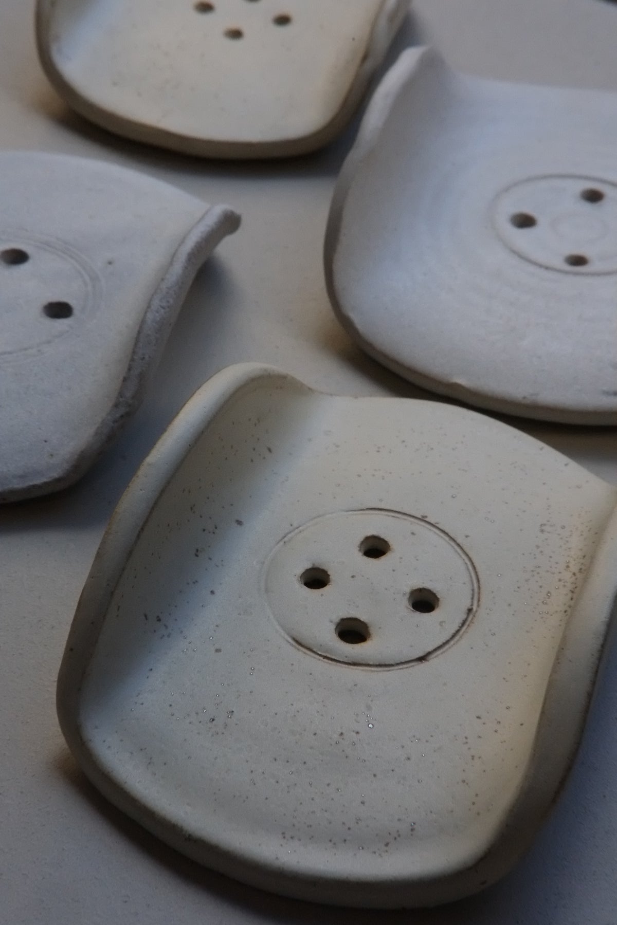 Four handmade ceramic soap dishes with a textured, speckled off-white glaze, arranged on a neutral light gray background.