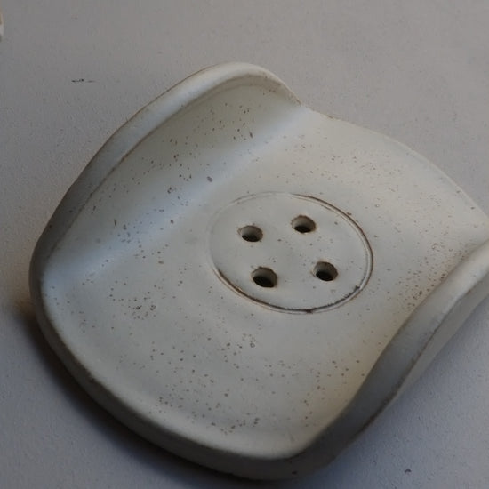 A white ceramic soap dish with a textured speckled finish, featuring a circular center with four drainage holes, photographed on a neutral grey background.