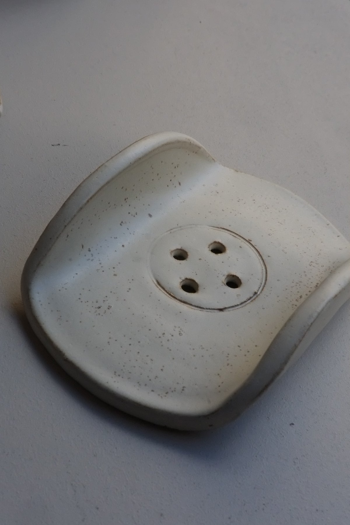 A white ceramic soap dish with a textured speckled finish, featuring a circular center with four drainage holes, photographed on a neutral grey background.