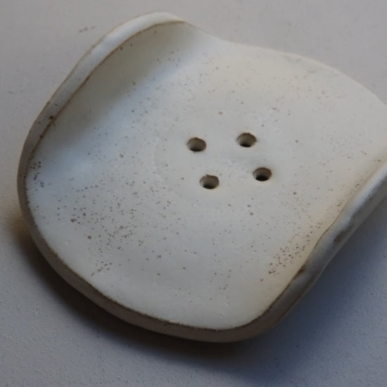 A white ceramic button with four holes and a slightly distressed, speckled finish, resting on a neutral light gray surface.