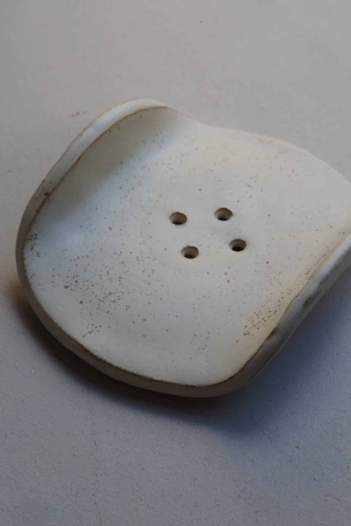 A white ceramic button with four holes and a slightly distressed, speckled finish, resting on a neutral light gray surface.