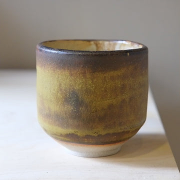 A handmade ceramic cup with a rustic glaze featuring a gradient of olive green, brown, and black, sitting on a light wooden surface against a neutral gray background.