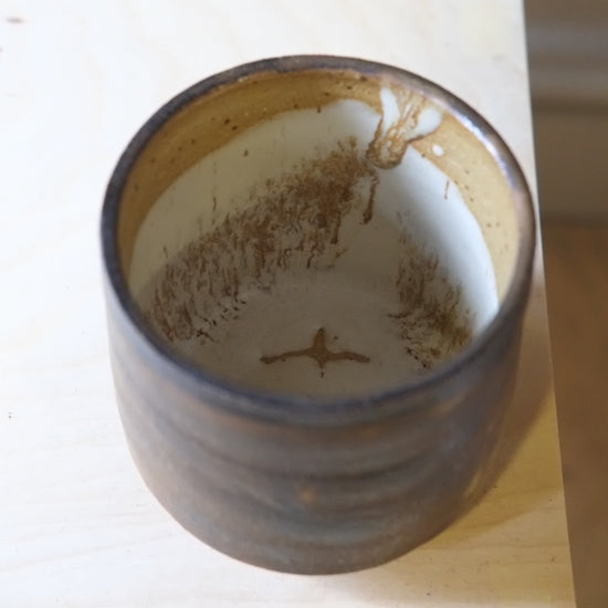 A handmade ceramic cup with a rustic finish, featuring a dark brown exterior and a warm beige interior with brown speckled details, shown on a light wood surface.