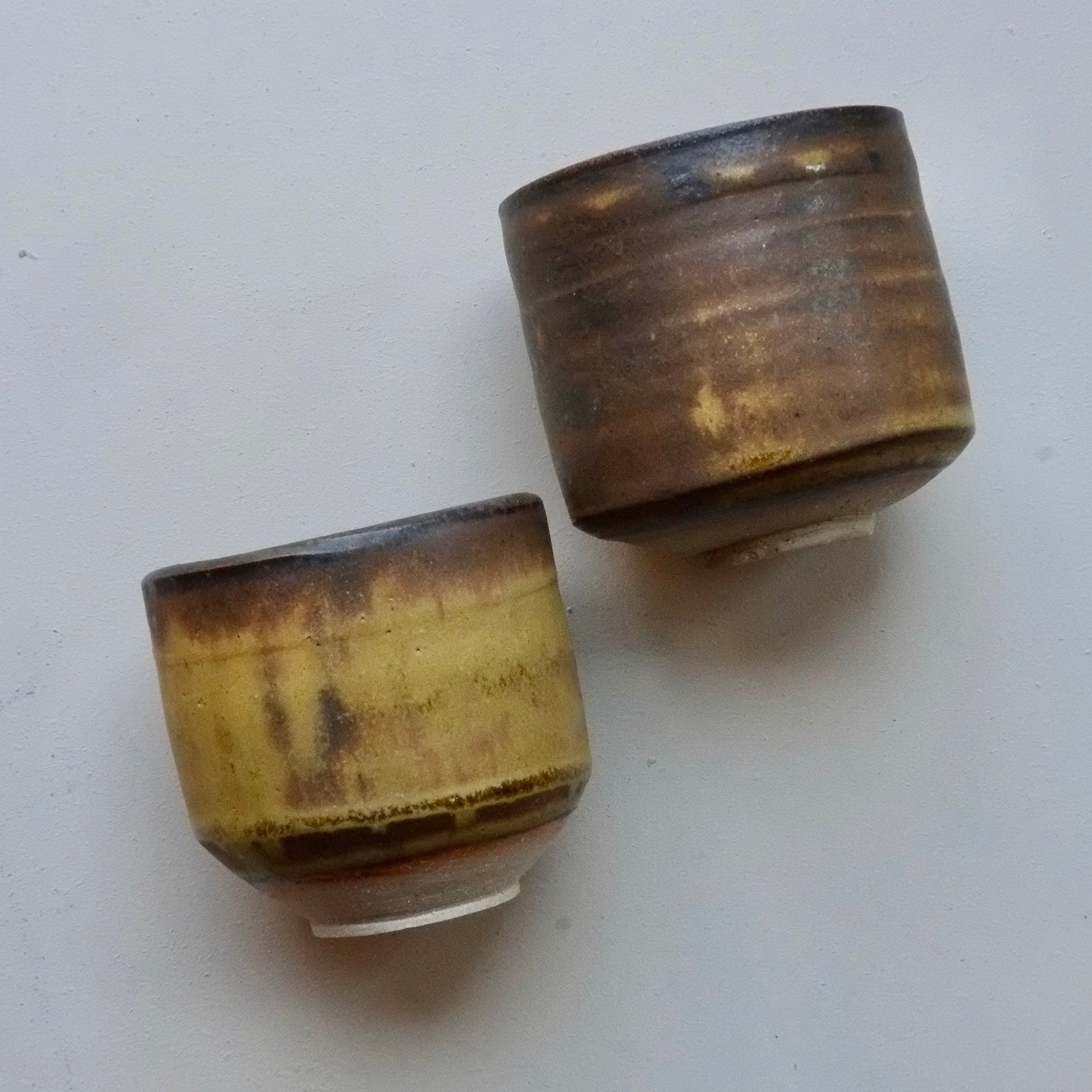 Two handmade ceramic cups with a rustic, hand-painted glaze in earthy brown, gold, and black tones, displayed on a light gray surface.