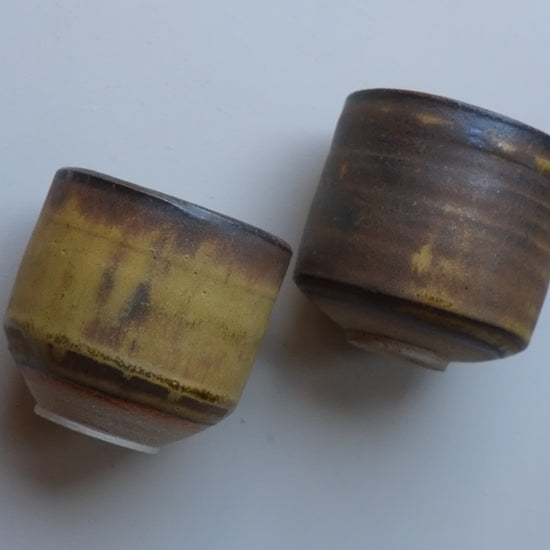 Two handmade ceramic cups with a rustic, glazed finish in brown and gold tones, displayed on a plain light gray surface.