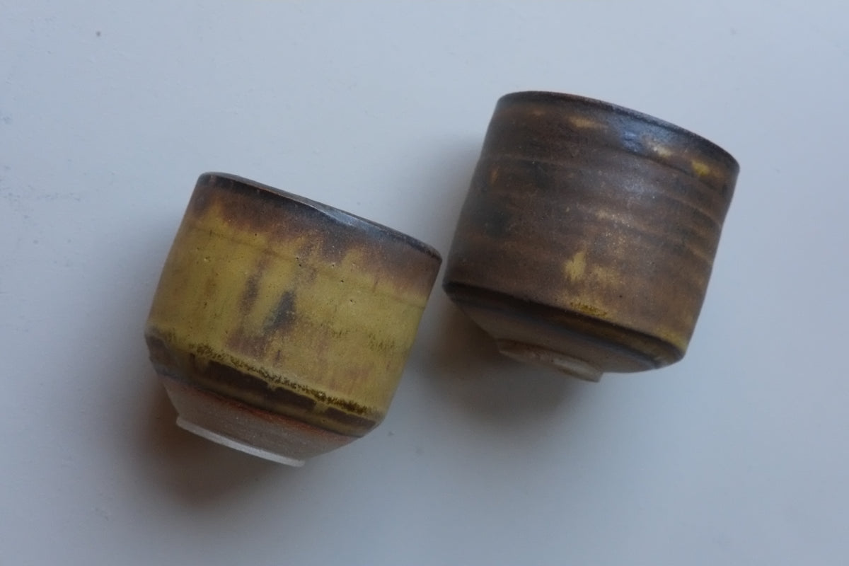 Two handmade ceramic cups with a rustic, glazed finish in brown and gold tones, displayed on a plain light gray surface.