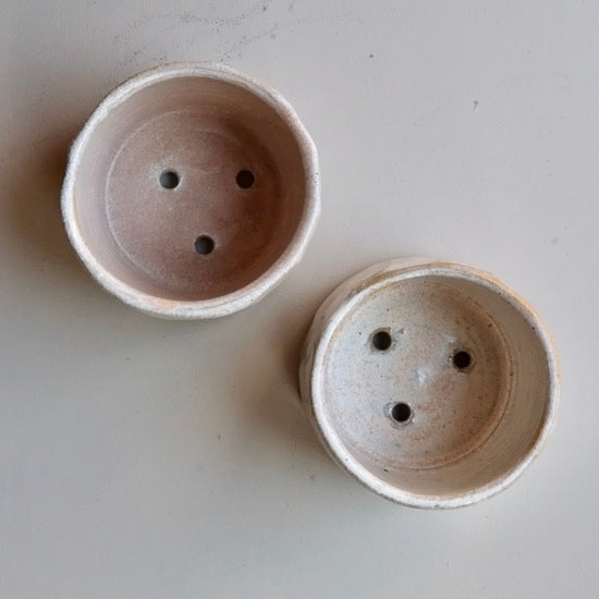 Two small round ceramic pots with drainage holes, shown from above on a light gray surface.