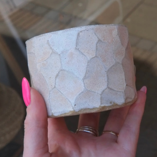 A hand with pink nail polish holds a small cylindrical planter pot with a textured, organic stone-like pattern in a light beige color.