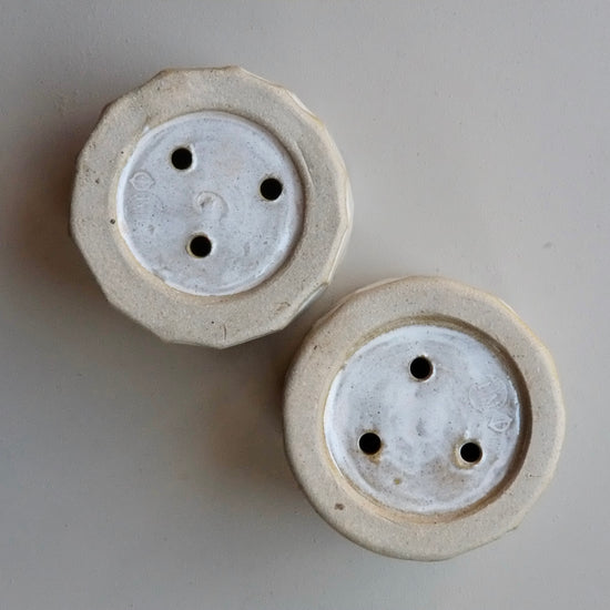 Two round ceramic pots shown from the top, featuring a textured off-white exterior and a glossy white interior with three holes each, set against a neutral gray background.