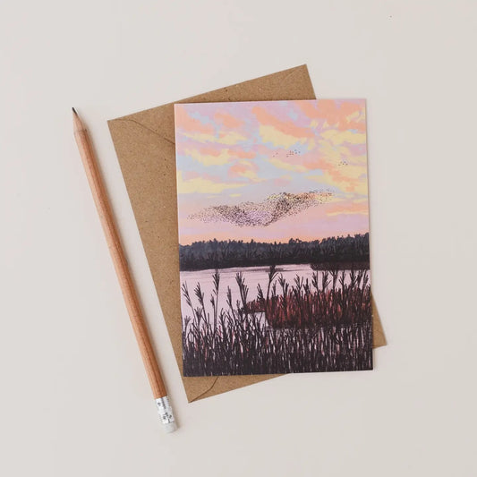 Greeting card with a sunset over water design, placed on a brown envelope with a pencil beside it.