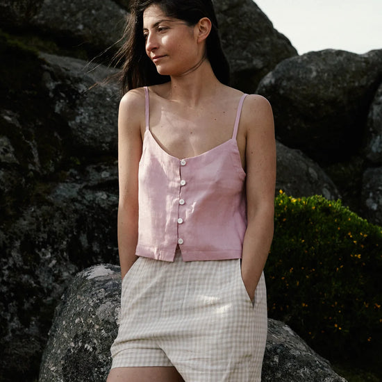Woman wearing a pale pink top with a button down front and thin straps and checkered shorts standing against a rocky background