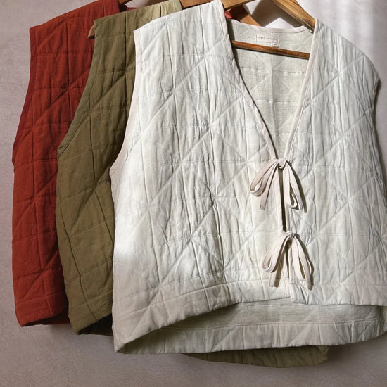 Three padded quilted sleeveless vests with two tie front fastening ribbons in cream, khaki and red hanging on wooden hangers in front of a grey background