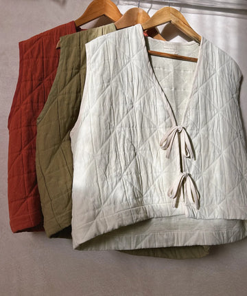 Three padded quilted sleeveless vests with two tie front fastening ribbons in cream, khaki and red hanging on wooden hangers in front of a grey background